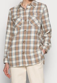 Vrouw in een lichtbeige, witte en blauwe geblokte blouse met lange mouwen en twee borstzakken, gecombineerd met lichte broek.