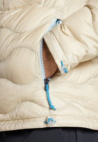 Beige quilted jacket with a smooth finish, featuring a blue zipper and drawstring accents, and an open cuff revealing skin underneath.