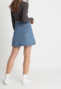 Jeans A-Linien-Rock mit hoher Taille, blauer Farbe, fransigem Saum und zwei Gesäßtaschen, kombiniert mit einem engen Langarmoberteil und weißen Stiefeln.