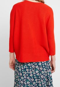 Czerwony, prążkowany sweter z trzema czwarte rękawami, luźny krój i okrągły dekolt, nałożony na spódnicę w kwiatowy wzór.