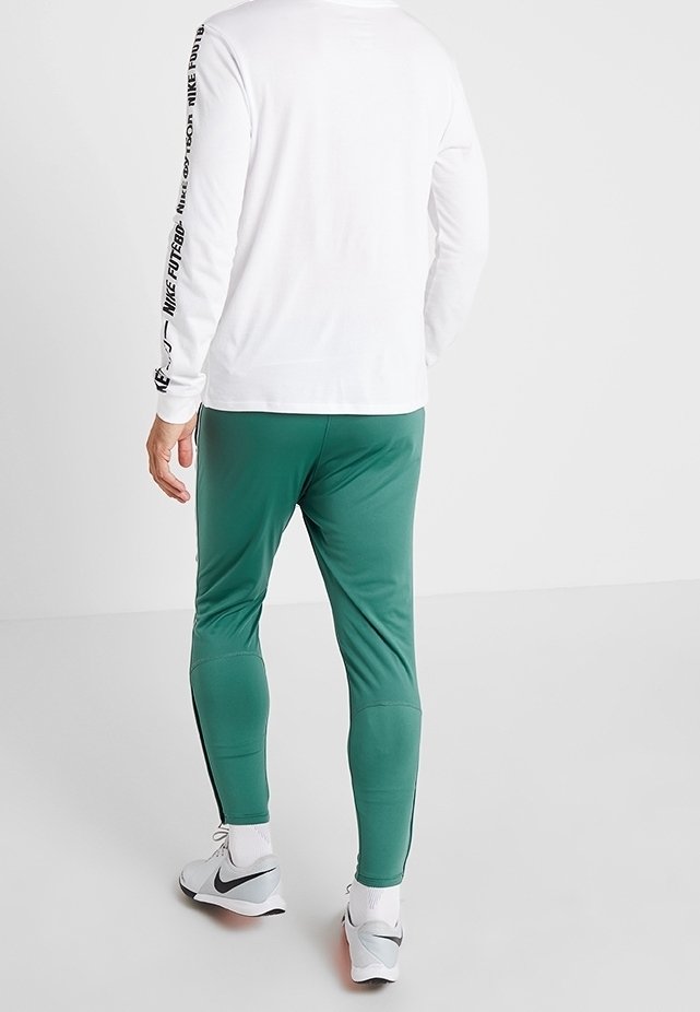 Person i vit långärmad skjorta med svart text på Ärmarna, gröna träningsbyxor, vita strumpor och vita Nike-skor, stående mot vit bakgrund.