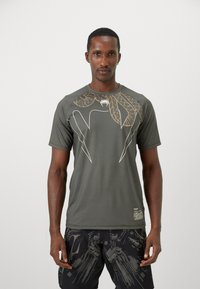 T-shirt de sport vert présentant un design graphique de feuilles dorées et un emblème d'araignée sur la poitrine. Fabriqué dans un tissu lisse et léger.