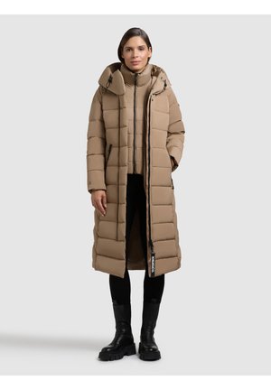 Piumino beige con un design lungo, dotato di cappuccio, chiusura a zip frontale e tasche laterali. Accenti neri vicino all'orlo, indossato sopra pantaloni e stivali neri.