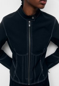 Veste noire avec surpiqûres blanches contrastantes, fermeture zippée à l'avant et col droit. Tissu lisse avec une silhouette ajustée et des coutures décoratives.