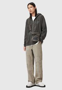 Dunkelgrauer Reißverschluss-Hoodie mit weißem Text, hellgraues T-Shirt, lockere sandfarbene Cargo-Hose mit sichtbaren Nähten, schwarz-weiße Sneaker, gestepptes Umhängetäschchen.