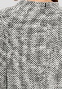 Tkaná látková bunda v černé a bílé barvě, vystupující s texturovaným houndstooth vzorem, s vysokým límcem a diskrétním zapínáním na zip na zadní straně.