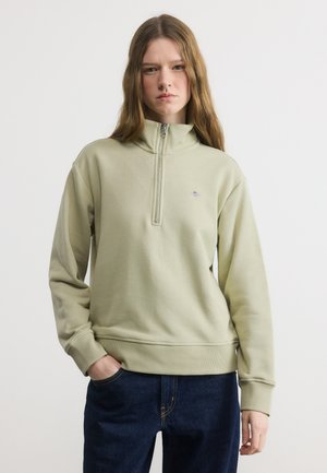 Young woman with long hair wearing light green quarter-zip sweatshirt and dark blue jeans, standing with one hand in pocket against white background.