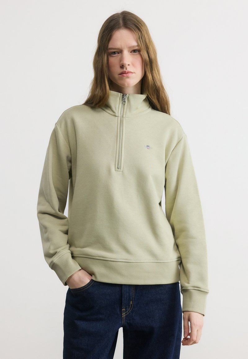 Νεαρή γυναίκα με μακριά μαλλιά, που φοράει ανοιχτοπράσινο φούτερ με φερμουάρ τέταρτου μήκους και σκούρα μπλε τζιν, στέκεται με το ένα χέρι στην τσέπη μπροστά σε λευκό φόντο.