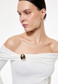 Weißes schulterfreies Oberteil mit drapiertem Stoff, ausgestattet mit einer glänzenden goldenen Brosche. Das Model hat glattes Haar und einen neutralen Hautton.