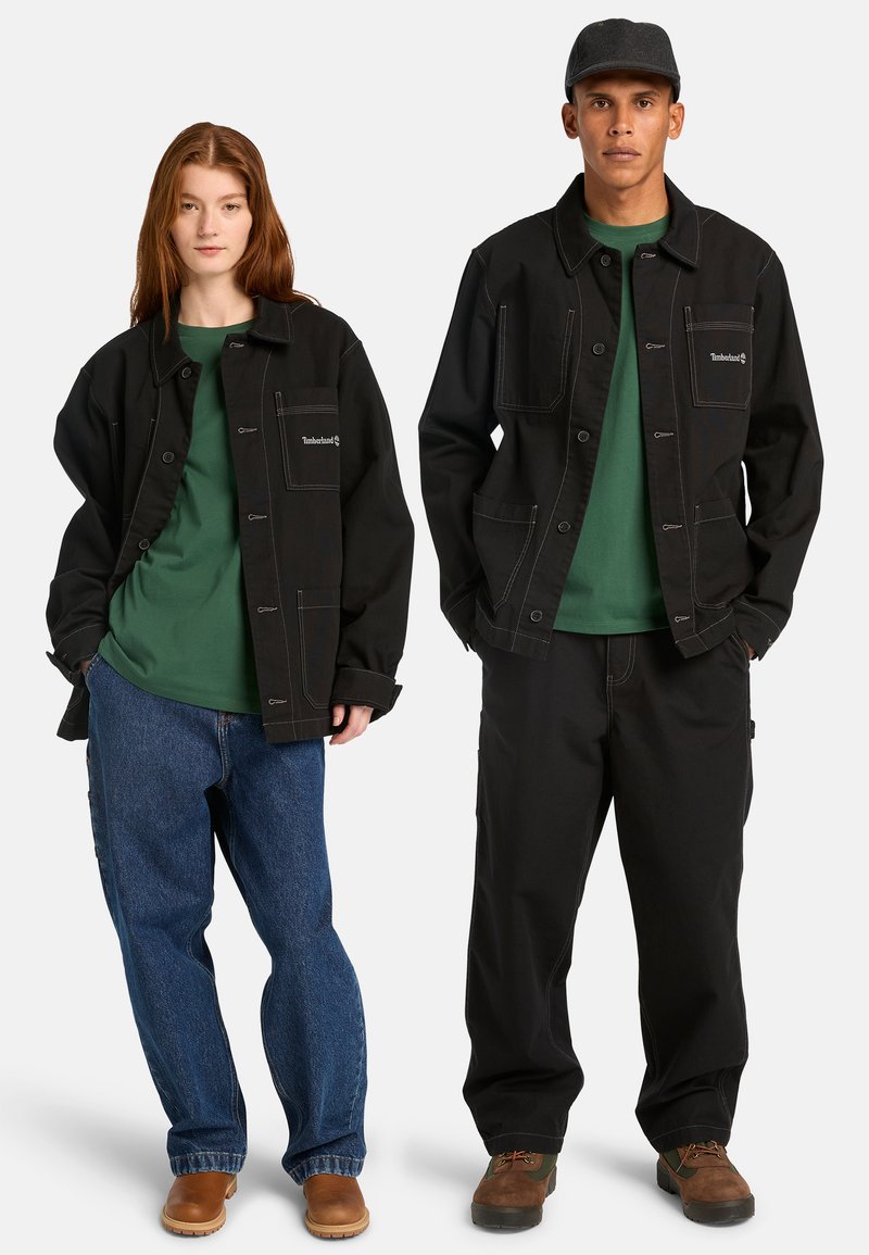 Vestes noires et durables avec plusieurs poches, t-shirts verts en dessous, associés à des jeans bleus et des pantalons noirs, complétés par des bottes marron.