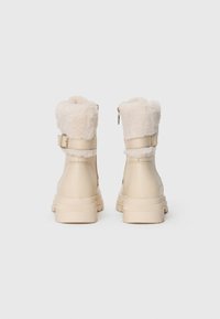 Beige ankelstøvler med en fluffy kremfarget krave og glidelås på siden, med teksturert såle og justerbar spenne. Myk materialfinish.