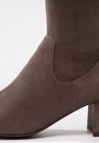 Bota corta de ante marrón con tacón cuadrado, que presenta una textura suave y un diseño ajustado, destacando los detalles de costura a lo largo de la costura.