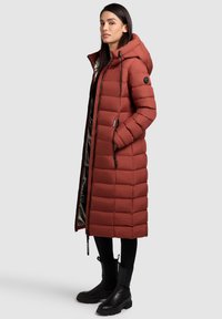 Long puffer coat in rust color, featuring a hood, zippered front, and contrasting inner lining. Made of quilted material with side pockets.