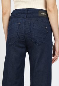 Dunkelblaue Denim-Jeans mit hoher Taille, ausgestattet mit zwei Gesäßtaschen und einem Lederetikett der Marke. Strukturierter Stoff mit leichtem Glanz.