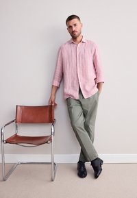 Ljusrosa randig skjorta, olivgröna byxor, svarta skor och en brun läderstol med kromade ben. Modellen lutar sig mot en vägg.