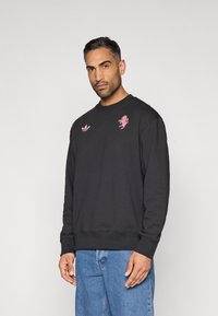 Felpa nera con scollo rotondo, caratterizzata da ricami rosa di un leone e dai loghi trefoil di Adidas. Realizzata in cotone, presenta polsini e orlo a coste.