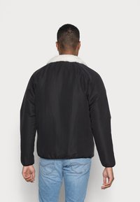 Sort jakke med en hvid kunstlæderkrave, der har en blød tekstur, elastiske manchetter og en afslappet pasform. Bæres over lyseblå jeans.