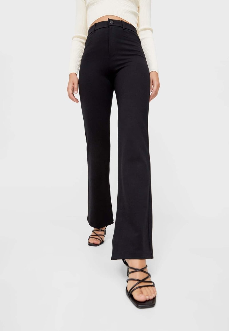 Femme portant un pantalon noir évasé taille haute et des sandales à talons noires à lanières, un haut blanc à manches longues raccourci, marchant vers l'avant.