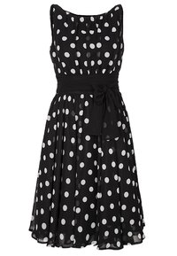 Robe noire sans manches à pois blancs, taille cintrée avec ceinture en tissu nouée, longueur au genou avec une jupe fluide.