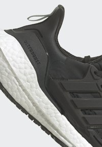 Sort atletisk sko med en tekstureret overdel, der har lysegrå detaljer og en hvid polstret sål. Viser "ULTRABOOST" branding.