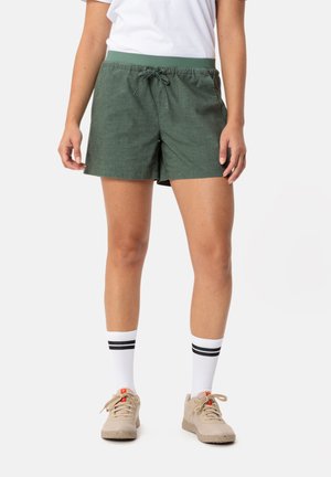 Shorts en lin verts avec une taille élastique et un cordon de serrage. Associés à des chaussettes blanches avec des rayures noires et des baskets beiges.