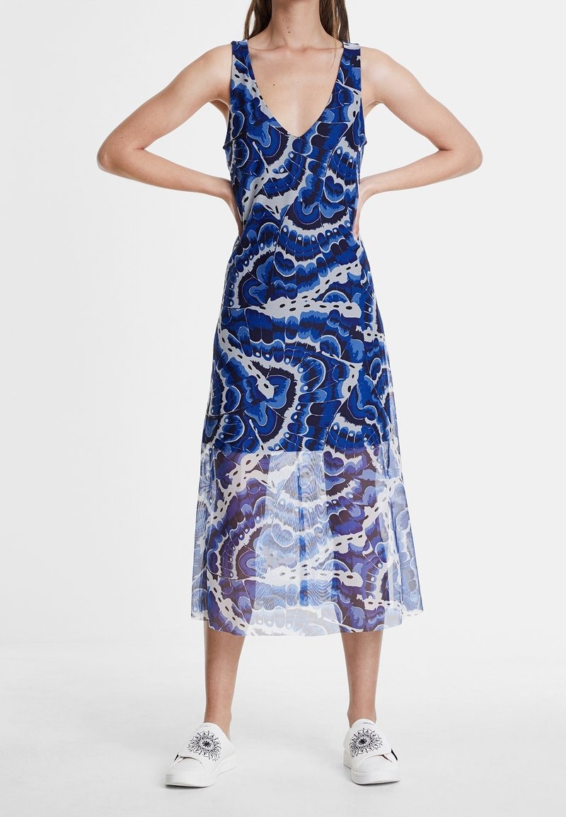 Robe midi sans manches à motifs bleus et blancs avec décolleté en V, portée avec des baskets blanches à enfiler ornées de motifs de soleils noirs.