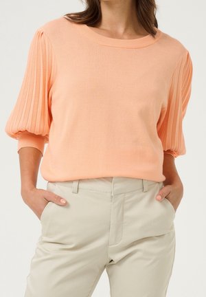 Vrouw die een perzikkleurige trui met korte pofmouwen en beige, hooggetailleerde broek draagt, met haar handen in de zakken, staande tegen een effen achtergrond.