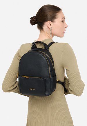 Naine, kellel on kuklapeas soeng, kannab beeźi värvi soonikkoes kleiti ja hõbedast ketist kõrvarõngaid, hoides käes musta nahast Liu Jo seljakotti kuldsete tõmblukkudega.