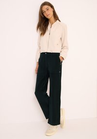 Vrouw met lang bruin haar in een lichtbeige zip-up jack, donkere wijde broek en witte sneakers, staand tegen een effen achtergrond.