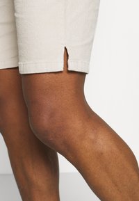 Primo piano delle cosce superiori di una persona che indossa pantaloni corti beige chiaro con una piccola fessura laterale, mostrando una pelle scura e una texture liscia.