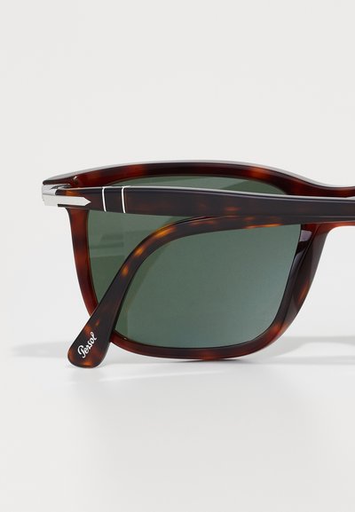 Óculos de sol em padrão tartaruga com lentes verdes, detalhes em metal nas hastes e uma armação curvada. Apresenta o logótipo da marca na haste.