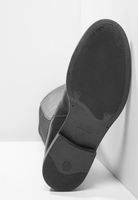 Suela de una bota de cuero negro inclinada contra dos paredes blancas, mostrando la talla 37 y la marca "Vagabond" estampada en la suela.