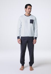 Camicia azzurro chiaro a maniche lunghe con tasca e polsini blu navy, abbinata a pantaloni jogger blu navy. Il materiale sembra morbido e casual.