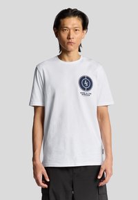 Hvid bomulds-t-shirt med et cirkulært marineblåt logo med teksten "LS" og "BORN IN THE BORDERS", korte ærmer og rund halsudskæring.