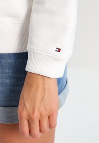 Witte sweatshirtmanchet van gestructureerde stof, met een klein marineblauw, rood en wit logo op de pols. Draagt hierbij denim shorts.
