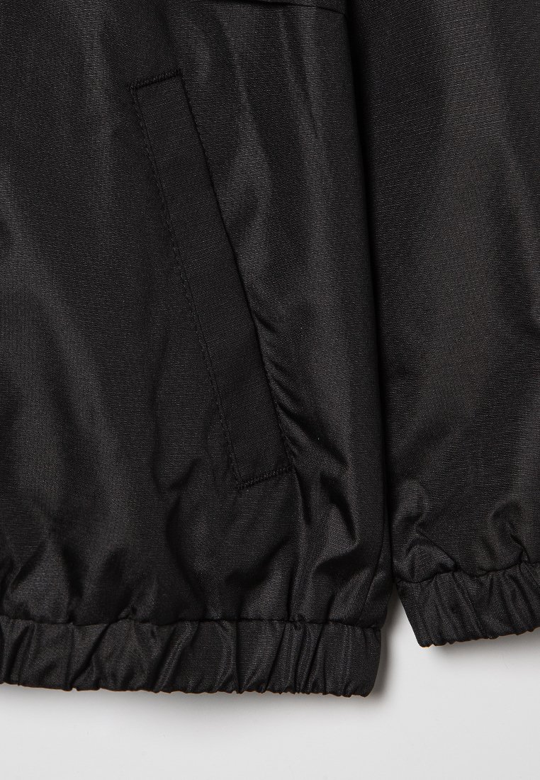 Veste en nylon noir avec une texture lisse, des poignets élastiques et une poche latérale. Présente un ourlet froncé et un design minimaliste.