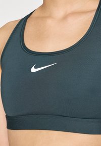 Tumši zaļš sporta krūšturis ar gludu tekstūru, platām siksnām un Apaļu dekoltē. Centrā priekšā ir baltais Nike logo.