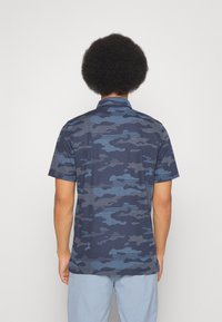 Koszula z krótkim rękawem w granatowym kamuflażu, z kołnierzykiem na guziki, fakturowaną tkaniną i luźnym krojem, widoczna od tyłu.