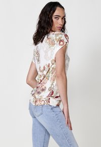 Haut à manches courtes à motifs floraux avec une base crème, détail texturé à l'ourlet, associé à un jean bleu clair avec des accents de perles sur le côté.