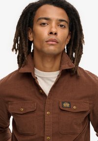 Bruine flanelle hemd met een spread collar, knoopsluiting, twee borstzakken en een logo-opdruk. De stof heeft een gestructureerde uitstraling.