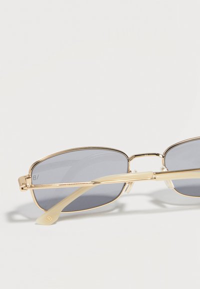 Ορθογώνια γυαλιά ηλίου με λεπτά χρυσά μεταλλικά πλαίσια και γκρι φακούς τους οποίους στηρίζουν σε μια επίπεδη λευκή επιφάνεια.