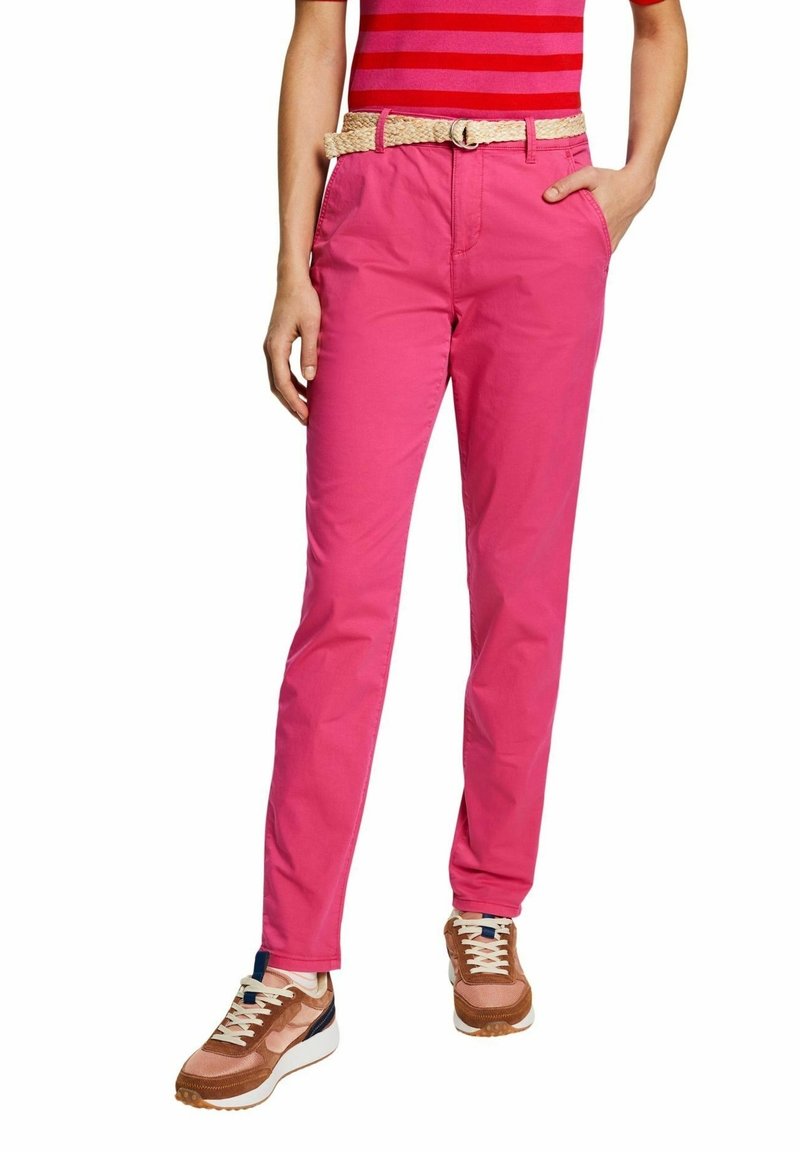 Personne portant un pantalon rose vif, une ceinture beige tressée, des baskets marron avec des lacets blancs et une chemise rayée rouge, les mains dans les poches.