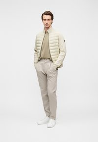 Επενδεδυμένο, ανοιχτό μπεζ μπουφάν με ψηλό γιακά πάνω από ένα απαλο πράσινο πουκάμισο, σε συνδυασμό με ανοιχτόχρωμο ριγέ παντελόνι και λευκά αθλητικά παπούτσια.