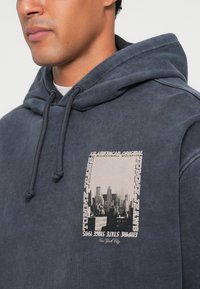 Hanorac bleu închis, fabricat dintr-un material moale, cu un grafic pe față ce înfățișează un orizont de oraș, împreună cu text, șnururi ajustabile și o croială lejeră.