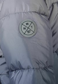 Ljuslila puffjacka med en glänsande, slät textur. Har en rund vit lapp med svarta geometriska mönster och texten "NMT 97".