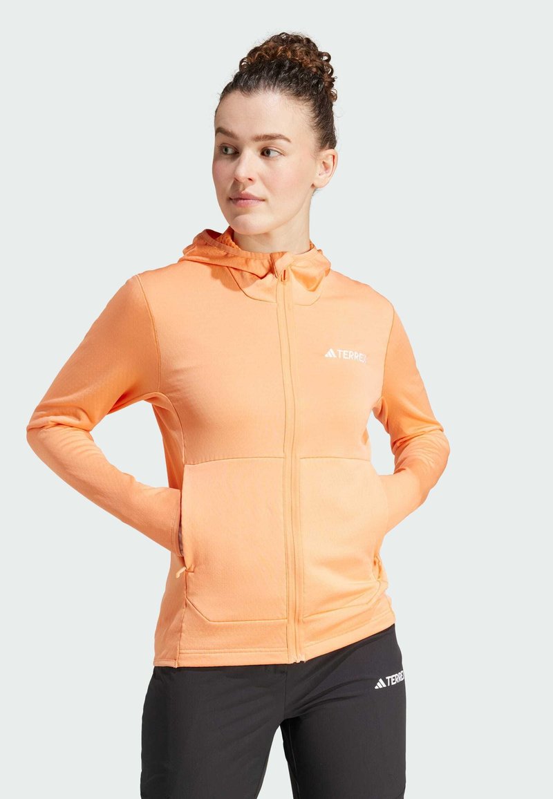 Sudadera ligera naranja con cremallera, capucha y dos bolsillos frontales, hecha de una tela suave. Presenta un logo en el pecho y un diseño ajustado.