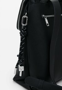 Bolso de mano de cuero negro con un acabado texturizado, cremallera plateada y un detalle en cuerda con patrón en blanco y negro con un detalle metálico.
