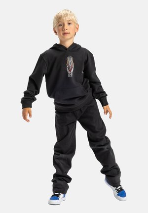 Zwarte hoodie met vlamgraphic, ontspannen pasvorm zwarte broek en sneakers met blauwe accenten. Model staat met ontspannen armen tegen een witte achtergrond.