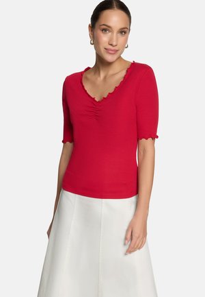 Frau trägt ein rotes Kurzarmshirt mit Rüschensaum am Ausschnitt und weißen Rock, steht vor einem schlichten hellen Hintergrund.