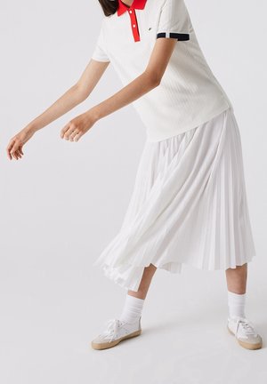 Personne en jupe plissée blanche, polo blanc avec col rouge, chaussettes blanches et baskets blanches, se penchant en avant avec les bras tendus.